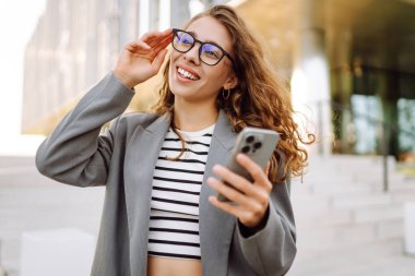 Şık giyinmiş, elinde telefonla modern binanın yakınında güneşli bir günün tadını çıkaran genç bir kadın. Şık bir kadın akıllı telefona bakıyor ve dışarıda gülümsüyor. Keyif ve rahatlama konsepti. Yaşam biçimi.