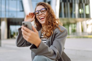 Şık giyinmiş, elinde telefonla modern binanın yakınında güneşli bir günün tadını çıkaran genç bir kadın. Şık bir kadın akıllı telefona bakıyor ve dışarıda gülümsüyor. Keyif ve rahatlama konsepti. Yaşam biçimi.