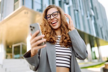 Şık giyinmiş, elinde telefonla modern binanın yakınında güneşli bir günün tadını çıkaran genç bir kadın. Şık bir kadın akıllı telefona bakıyor ve dışarıda gülümsüyor. Keyif ve rahatlama konsepti. Yaşam biçimi.