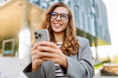 Şık giyinmiş, elinde telefonla modern binanın yakınında güneşli bir günün tadını çıkaran genç bir kadın. Şık bir kadın akıllı telefona bakıyor ve dışarıda gülümsüyor. Keyif ve rahatlama konsepti. Yaşam biçimi.