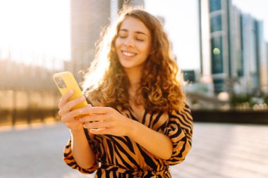 Genç bir kadının şehir merkezinin arka planında tuttuğu sarı bir telefonun yakın çekimi. Elbise giymiş güzel bir kadın elinde telefonla mesajlaşıyor. Kentsel çevre kavramı.