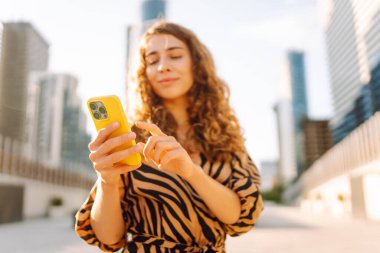 Genç bir kadının şehir merkezinin arka planında tuttuğu sarı bir telefonun yakın çekimi. Elbise giymiş güzel bir kadın elinde telefonla mesajlaşıyor. Kentsel çevre kavramı.