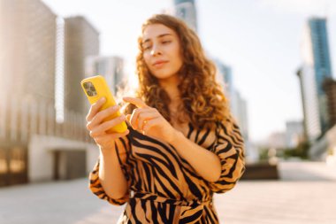 Genç bir kadının şehir merkezinin arka planında tuttuğu sarı bir telefonun yakın çekimi. Elbise giymiş güzel bir kadın elinde telefonla mesajlaşıyor. Kentsel çevre kavramı.