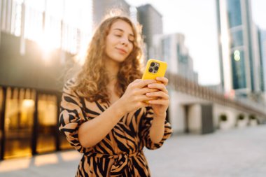 Genç bir kadının şehir merkezinin arka planında tuttuğu sarı bir telefonun yakın çekimi. Elbise giymiş güzel bir kadın elinde telefonla mesajlaşıyor. Kentsel çevre kavramı.