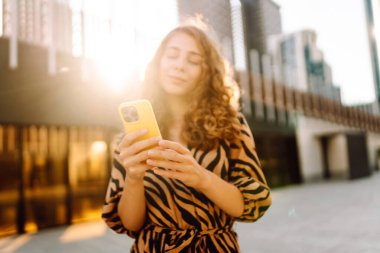 Genç bir kadının şehir merkezinin arka planında tuttuğu sarı bir telefonun yakın çekimi. Elbise giymiş güzel bir kadın elinde telefonla mesajlaşıyor. Kentsel çevre kavramı.