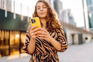 Genç bir kadının şehir merkezinin arka planında tuttuğu sarı bir telefonun yakın çekimi. Elbise giymiş güzel bir kadın elinde telefonla mesajlaşıyor. Kentsel çevre kavramı.