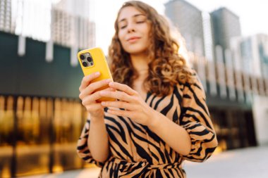 Genç bir kadının şehir merkezinin arka planında tuttuğu sarı bir telefonun yakın çekimi. Elbise giymiş güzel bir kadın elinde telefonla mesajlaşıyor. Kentsel çevre kavramı.