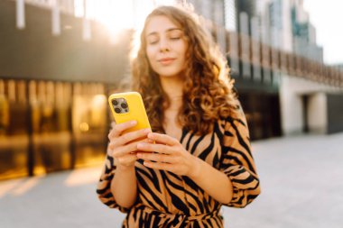 Genç bir kadının şehir merkezinin arka planında tuttuğu sarı bir telefonun yakın çekimi. Elbise giymiş güzel bir kadın elinde telefonla mesajlaşıyor. Kentsel çevre kavramı.