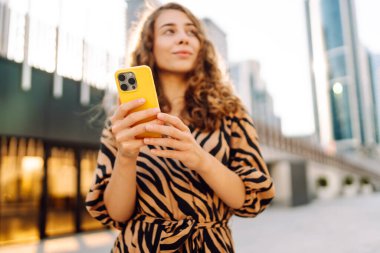 Genç bir kadının şehir merkezinin arka planında tuttuğu sarı bir telefonun yakın çekimi. Elbise giymiş güzel bir kadın elinde telefonla mesajlaşıyor. Kentsel çevre kavramı.