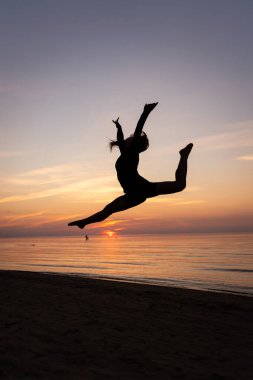 Güzel bir kadının silueti, deniz manzarasına karşı zarif bir poz veriyor. Genç bir jimnastikçi gün batımında çeşitli spor unsurları sergiler. Spor, güzellik ve rahatlama kavramı.