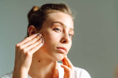 Genç bir kadının, aynaya bakarken yüzünü beyaz pamukla silişi. Genç bir kadın sıcak odada yansımayı ve yüzlere dokunmayı sever. Güzellik ve doğallık kavramı.