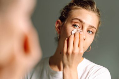 Genç bir kadının, aynaya bakarken yüzünü beyaz pamukla silişi. Genç bir kadın sıcak odada yansımayı ve yüzlere dokunmayı sever. Güzellik ve doğallık kavramı.