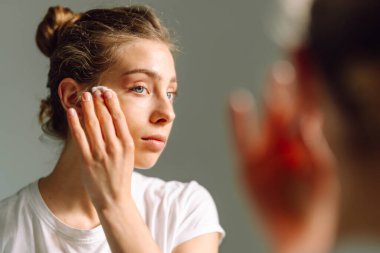 Genç bir kadının, aynaya bakarken yüzünü beyaz pamukla silişi. Genç bir kadın sıcak odada yansımayı ve yüzlere dokunmayı sever. Güzellik ve doğallık kavramı.
