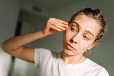 Genç bir kadının, aynaya bakarken yüzünü beyaz pamukla silişi. Genç bir kadın sıcak odada yansımayı ve yüzlere dokunmayı sever. Güzellik ve doğallık kavramı.