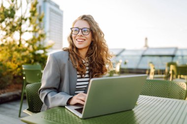 Dizüstü bilgisayarı olan gülümseyen bir kadın şehrin manzarasının tadını çıkarıyor. Gün batımında dışarıda çalışan güzel bir kadın serbest yazar. İş, eğitim ve eğlence konsepti. Uzak çalışma.