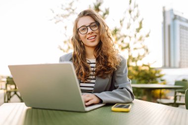 Dizüstü bilgisayarı olan gülümseyen bir kadın şehrin manzarasının tadını çıkarıyor. Gün batımında dışarıda çalışan güzel bir kadın serbest yazar. İş, eğitim ve eğlence konsepti. Uzak çalışma.