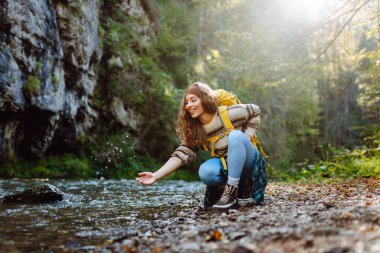 Yürüyüş yapan parlak sarı sırt çantalı güzel bir kadın dağ nehrine elleriyle dokunuyor ve yolculuğun tadını çıkarıyor. Genç bayan gezgin dağlarda yürüyüş parkurunda. Etkin yaşam biçimi.
