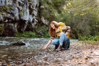 Yürüyüş yapan parlak sarı sırt çantalı güzel bir kadın dağ nehrine elleriyle dokunuyor ve yolculuğun tadını çıkarıyor. Genç bayan gezgin dağlarda yürüyüş parkurunda. Etkin yaşam biçimi.