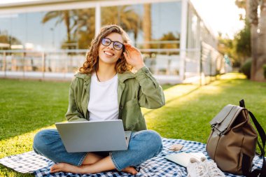 Dizüstü bilgisayarı olan genç bir kadın palmiye ağacının altındaki çimlerde havanın tadını çıkarıyor. Neşeli kadın serbest çalışan ya da dışarıda okuyan. Rahatlama kavramı, uzak çalışma.