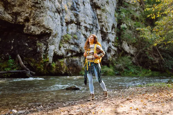 Yürüyüş yapan parlak sarı sırt çantalı güzel bir kadın dağ nehrine elleriyle dokunuyor ve yolculuğun tadını çıkarıyor. Genç bayan gezgin dağlarda yürüyüş parkurunda. Etkin yaşam biçimi.