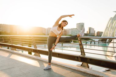 Spor kıyafetli genç bir kadın köprü manzaralı şehirde egzersiz yapıyor. Gün batımı ışınlı atletik kadın dışarıda egzersiz yapıyor. Spor konsepti. Etkin yaşam biçimi.