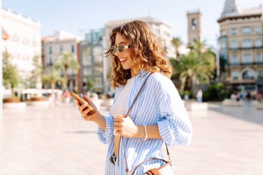 Güneş gözlüklü, elinde telefon tutan neşeli bir genç kadın güneşli sokaklarda yürüyor. Telefonu olan mutlu bir turist güneşli bir günde yeni bir mimarinin tadını çıkarır. Turizm ve keyif konsepti.