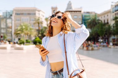 Güneş gözlüklü, elinde telefon tutan neşeli bir genç kadın güneşli sokaklarda yürüyor. Telefonu olan mutlu bir turist güneşli bir günde yeni bir mimarinin tadını çıkarır. Turizm ve keyif konsepti.