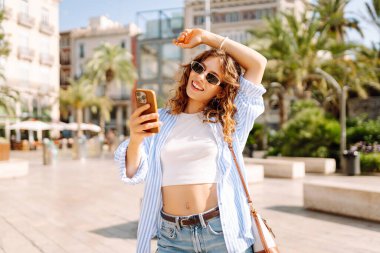 Güneş gözlüklü, elinde telefon tutan neşeli bir genç kadın güneşli sokaklarda yürüyor. Telefonu olan mutlu bir turist güneşli bir günde yeni bir mimarinin tadını çıkarır. Turizm ve keyif konsepti.
