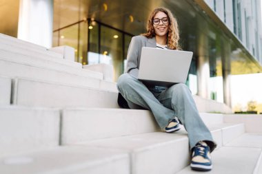 Modern binanın arka planında dizüstü bilgisayarı olan şık bir kadın. Gözlüklü bir iş kadını dışarıda dizüstü bilgisayarda çalışıyor. Bağımsızlık, iş ve yıkım kavramı.