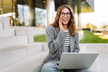 Modern binanın arka planında dizüstü bilgisayarı olan şık bir kadın. Gözlüklü bir iş kadını dışarıda dizüstü bilgisayarda çalışıyor. Bağımsızlık, iş ve yıkım kavramı.