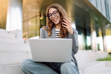 Modern binanın arka planında dizüstü bilgisayarı olan şık bir kadın. Gözlüklü bir iş kadını dışarıda dizüstü bilgisayarda çalışıyor. Bağımsızlık, iş ve yıkım kavramı.