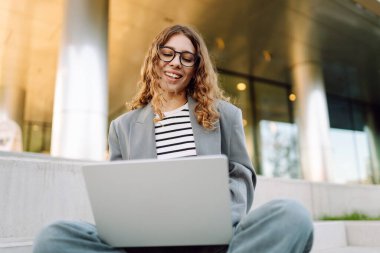 Modern binanın arka planında dizüstü bilgisayarı olan şık bir kadın. Gözlüklü bir iş kadını dışarıda dizüstü bilgisayarda çalışıyor. Bağımsızlık, iş ve yıkım kavramı.