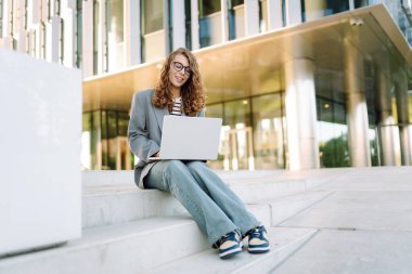Modern binanın arka planında dizüstü bilgisayarı olan şık bir kadın. Gözlüklü bir iş kadını dışarıda dizüstü bilgisayarda çalışıyor. Bağımsızlık, iş ve yıkım kavramı.