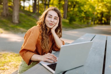 Sunny Park 'taki masada oturan dizüstü bilgisayarı ve telefonu olan neşeli bir kadın. Genç bayan serbest çalışan dışarıda dizüstü bilgisayarla çalışıyor. Uzak çalışma, doğa ve günlük tutma kavramı.