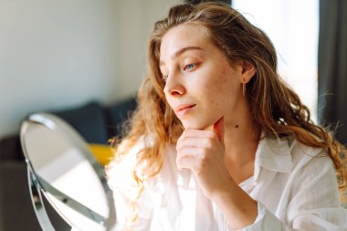 Genç bir kadının portresi, sıcacık bir odada elleri ve yüzlerine dokunuyor. Sivilceli bir kadın gün batımında küçük aynaya bakarken sivilceleri inceliyor. Kozmetik ve dermatoloji kavramı.
