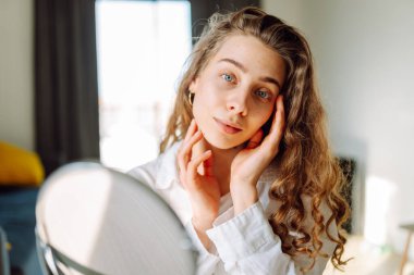 Genç bir kadının portresi, sıcacık bir odada elleri ve yüzlerine dokunuyor. Sivilceli bir kadın gün batımında küçük aynaya bakarken sivilceleri inceliyor. Kozmetik ve dermatoloji kavramı.