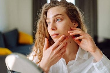 Genç bir kadının portresi, sıcacık bir odada elleri ve yüzlerine dokunuyor. Sivilceli bir kadın gün batımında küçük aynaya bakarken sivilceleri inceliyor. Kozmetik ve dermatoloji kavramı.