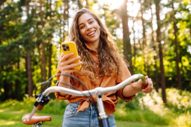 Telefonu ve bisikleti olan hoş bir kadın dışarıda eğleniyor. Güneşli parkta bisiklete binen ve telefonla mesajlaşan güzel bir kadın. Hafta sonu konsepti. Etkin yaşam biçimi.