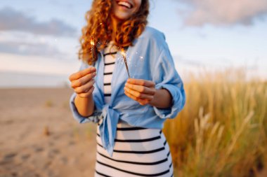 Gün batımında sahilde elinde maytap tutan genç bir kadın. Güzel bir kadın dışarıda eğleniyor ve bayramı kutluyor. Rahatlama kavramı, eğlence.