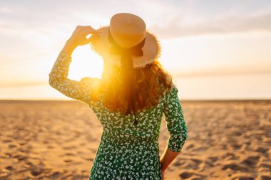 Şapkalı mutlu kadın gün batımını seyrediyor. Gün batımında sahilde duruyor. Genç bayan hava ve deniz manzarasının tadını çıkarıyor. Zevk ve özgürlük kavramı.