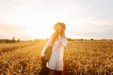 Beyaz elbiseli neşeli kadın altın buğday tarlasında poz veriyor. Genç bayan eğleniyor ve açık havada güneşli havanın tadını çıkarıyor. Doğa ve keyif kavramı.