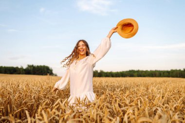 Beyaz elbiseli neşeli kadın altın buğday tarlasında poz veriyor. Genç bayan eğleniyor ve açık havada güneşli havanın tadını çıkarıyor. Doğa ve keyif kavramı.