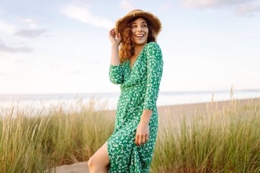 Gün batımında kumlu sahilde yürüyen mutlu kadın. Şapkalı genç turist dinleniyor ve havanın tadını çıkarıyor. Güzellik, eğlence konsepti. Etkin yaşam biçimi.