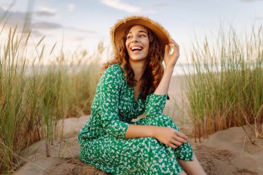 Gün batımında kumlu sahilde yürüyen mutlu kadın. Şapkalı genç turist dinleniyor ve havanın tadını çıkarıyor. Güzellik, eğlence konsepti. Etkin yaşam biçimi.