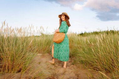 Gün batımında kumlu sahilde yürüyen mutlu kadın. Şapkalı genç turist dinleniyor ve havanın tadını çıkarıyor. Güzellik, eğlence konsepti. Etkin yaşam biçimi.