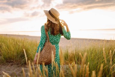 Gün batımında kumlu sahilde yürüyen mutlu kadın. Şapkalı genç turist dinleniyor ve havanın tadını çıkarıyor. Güzellik, eğlence konsepti. Etkin yaşam biçimi.