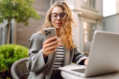 Genç bir kadın elinde telefon ve dizüstü bilgisayarla verandadaki bir masada oturuyor. Bir iş kadını kahve içer ve dışarıda dizüstü bilgisayarla çalışır. Serbest ve kahve molası kavramı.