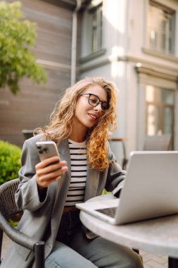 Genç bir kadın elinde telefon ve dizüstü bilgisayarla verandadaki bir masada oturuyor. Bir iş kadını kahve içer ve dışarıda dizüstü bilgisayarla çalışır. Serbest ve kahve molası kavramı.