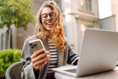 Genç bir kadın elinde telefon ve dizüstü bilgisayarla verandadaki bir masada oturuyor. Bir iş kadını kahve içer ve dışarıda dizüstü bilgisayarla çalışır. Serbest ve kahve molası kavramı.
