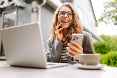Genç bir kadın elinde telefon ve dizüstü bilgisayarla verandadaki bir masada oturuyor. Bir iş kadını kahve içer ve dışarıda dizüstü bilgisayarla çalışır. Serbest ve kahve molası kavramı.